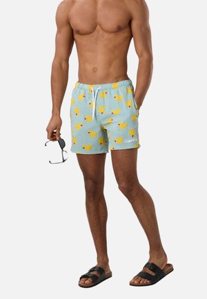 Homme torse nu portant un short de bain bleu clair avec un motif de canards en caoutchouc jaunes, des sandales noires, tenant des lunettes de soleil noires dans la main gauche.