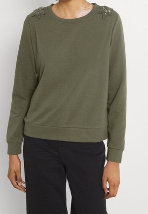 Personne portant un haut à manches longues vert olive avec des embellissements de feuilles perlées sur les épaules et un pantalon noir sur un fond uni.