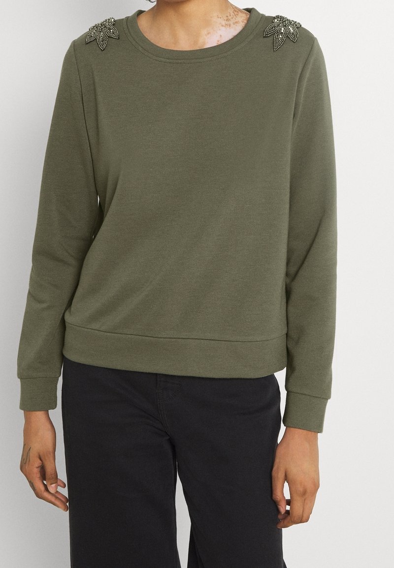 Personne portant un haut à manches longues vert olive avec des embellissements de feuilles perlées sur les épaules et un pantalon noir sur un fond uni.