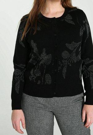 Femme portant un cardigan noir boutonné avec des motifs floraux argentés et un pantalon gris texturé, debout devant un fond uni.