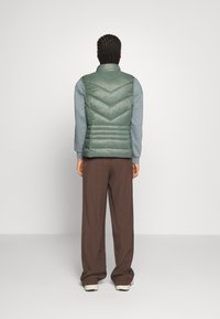Grüner Puffer-Weste mit Zickzacksteppung, hohem Kragen und horizontalen Abschnitten. Getragen über einem hellblauen Langarmshirt und braunen Hosen.