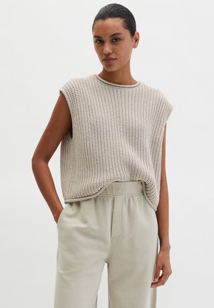 Woman wearing sleeveless knitted beige top tucked into high-waisted light beige pants with hands in pockets against white background.