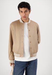 Beige bomberjakke med en glatt tekstur, stripete krage og sølvfargede trykknapper. Brukt over en hvit skjorte, kombinert med blå jeans.