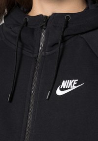 Černá mikina na zip z měkké látky, s vysokým límcem, šňůrkami a bílým logem Nike na levé straně hrudi.