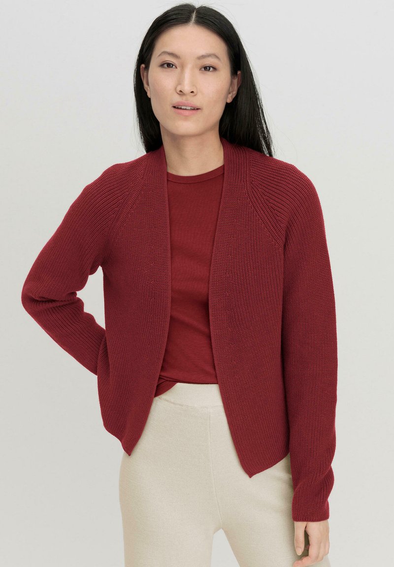 Rote Strickjacke mit Schalkragen und offenem Vorderteil; gerippte Textur, lange Ärmel, kombiniert mit einem roten Oberteil und hellen Hosen.