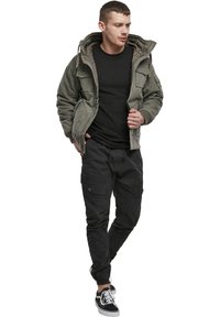 Olivegrüner, gepolsterter Jacke mit einer mit Kunstpelz gefütterten Kapuze, schwarzes Langarmshirt, dunkle Cargo-Hosen und schwarze Sneakers mit weißen Akzenten.
