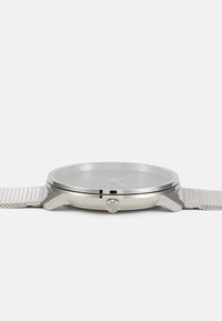 Reloj de acero inoxidable plateado con un diseño minimalista, que presenta una correa de malla, una tapa de cristal mineral plano y un dial sencillo con tres agujas.