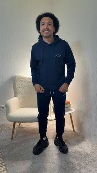 Man with curly hair wearing a navy hoodie and joggers stands smiling in a carpeted room with a white armchair and small table with books.