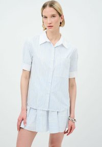Camicia button-up a righe verticali blu chiaro e bianche, maniche corte, colletto a punta, taschino sul lato sinistro del petto, abbinata a una gonna plissettata.