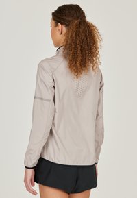 Lightweight, beige athletic jacket with a stand collar, elastic cuffs, and reflective accents. Features a perforated back for ventilation.