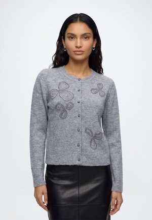 Femme portant un cardigan gris avec des motifs de papillons en perles et une jupe en cuir noire, debout devant un fond blanc uni.