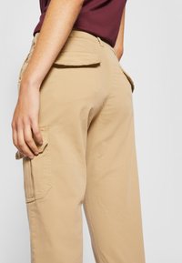 Beige cargo bukser med en afslappet pasform, der har sidelommer og en glat bomuldsstruktur. Taljen er synlig og komplementerer designet.