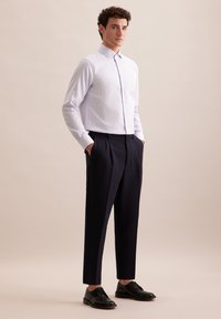 Lichtblauw gekleed overhemd met lange mouwen, aansluitend, tot aan de kraag geknoopt. Gecombineerd met marineblauwe wijde broek en zwarte veterschoenen.