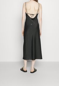 Svart satin-sleepekjole med tunna band och låg rygg, med en knyt-detalj. Tyget har en slät, blank yta.