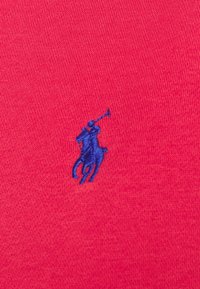 Tessuto di cotone rosso con una superficie liscia, caratterizzato da un piccolo logo ricamato blu di un cavallo e un cavaliere sul lato sinistro.
