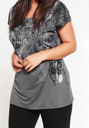 Vrouw draagt een grijs kortarmig shirt met zwart-witte abstracte patronen en strass details, gecombineerd met een zwarte broek.