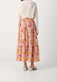 Jupe maxi fleurie avec un design superposé dans des teintes de rose, orange et jaune ; tissu léger ; taille élastique ; associée à un pull crème et des sandales.