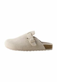 Zuecos de ante beige con puntera redondeada, una correa lateral con hebilla y suela de goma texturizada. Presentan un brillo sutil y un acento similar al corcho.