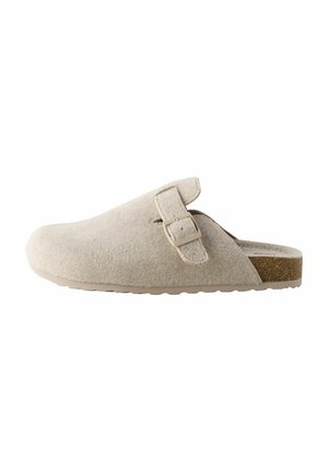 Zuecos de ante beige con puntera redondeada, una correa lateral con hebilla y suela de goma texturizada. Presentan un brillo sutil y un acento similar al corcho.