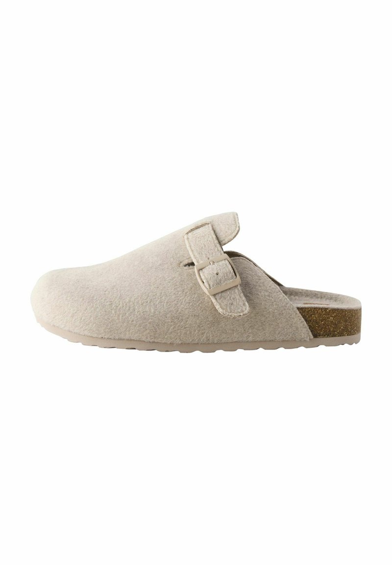 Zuecos de ante beige con puntera redondeada, una correa lateral con hebilla y suela de goma texturizada. Presentan un brillo sutil y un acento similar al corcho.