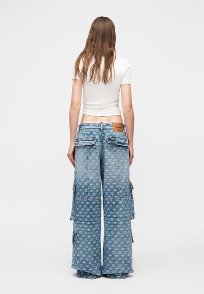 Femme aux longs cheveux bruns portant un crop top blanc à manches courtes et un pantalon cargo en denim bleu à jambes larges avec un motif intégral et des poches à rabat, vue de dos.