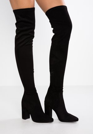 Femme portant des cuissardes en daim noir à talons carrés, debout devant un fond clair uni.