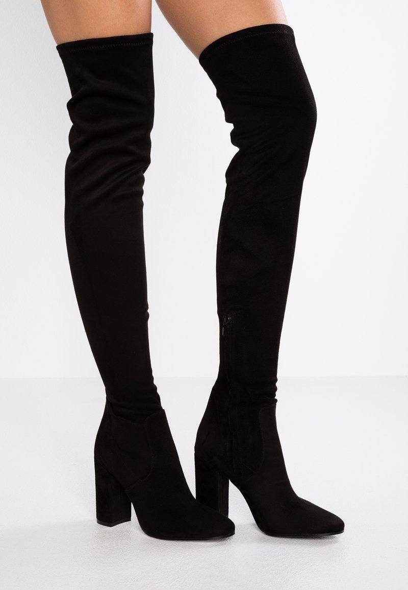 Femme portant des cuissardes en daim noir à talons carrés, debout devant un fond clair uni.