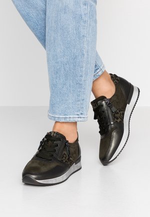 Personne portant des baskets noires et vert foncé avec des accents imprimé léopard et des fermetures éclair, assorties à un jean court bleu clair.