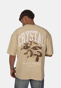 Béžové oversized tričko z m ěkké bavlny s bílým grafickým textem "CRYSTAL" a hnědou větvičkou citronu na zadní straně, krátké rukávy.