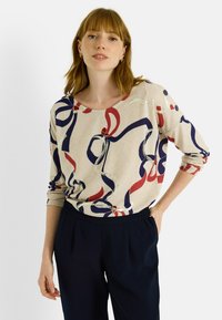 Haut à manches longues beige clair avec un motif de rubans multicolores en marine, rouge et accents métalliques. Encolure arrondie et détails de boutons sur l'épaule.