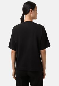 Person med mørkt hår sat tilbage iført en simpel sort kortærmet T-shirt og sorte bukser, vendt væk mod en hvid baggrund.