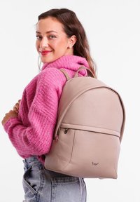 Light pink leather backpack with a rounded shape, top handle, and adjustable straps. Features a front zip pocket and subtle logo detail.