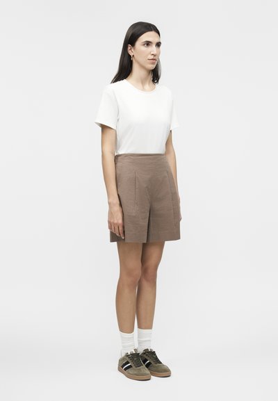 Frau mit langen dunklen Haaren steht nach rechts gewandt und trägt ein weißes kurzärmliges Hemd, braune strukturierte Shorts, weiße Socken und braune Turnschuhe.