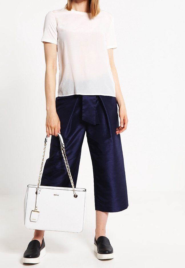 Femme portant un haut blanc à manches courtes, pantalon large cropped bleu marine, chaussures noires slip-on, tenant un sac à main blanc avec des anses en chaîne dorée.
