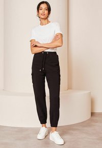 Sorte bomuldsbukser med snørebånd, sidelommer og elastiske manchetter, parret med en simpel hvid t-shirt og hvide sneakers på en neutral baggrund.