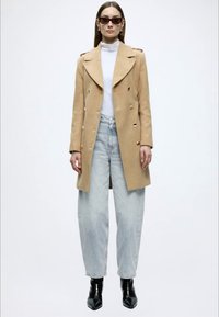 Beež topeltreaga mantel kuldsete nuppudega, kantud valge kaelusega kampsuni peal, kombineeritud helesiniste vabalt istuvate teksade ja mustade pahkluu saabastega.