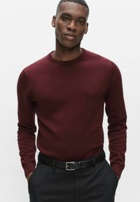 Bordeauxrode trui met ronde hals, lange mouwen, zachte textuur, gecombineerd met donkere broek en een zwarte riem met een zilveren gesp.