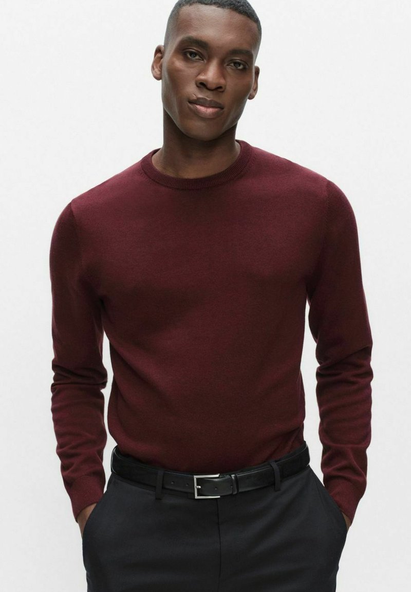 Bordeauxrode trui met ronde hals, lange mouwen, zachte textuur, gecombineerd met donkere broek en een zwarte riem met een zilveren gesp.