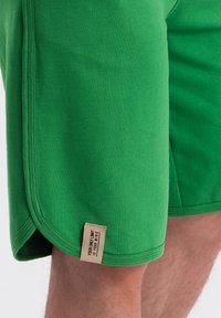 Shorts en coton vert avec une texture lisse, dotés d'une fente sur le côté et d'une étiquette beige avec du texte au ourlet. Design et coupe simples.