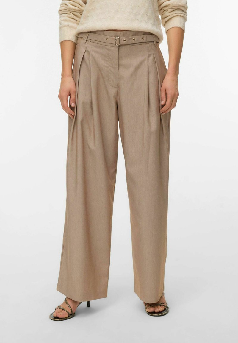Pantalon beige ample à taille haute avec des plis et une ceinture assortie, porté avec un pull crème et des sandales à talons ouvertes.