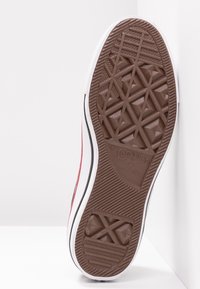 Semelle à motifs bruns d'une basket blanche avec des rayures noires et rouges, affichant le texte "Converse All Star" et le logo étoile.