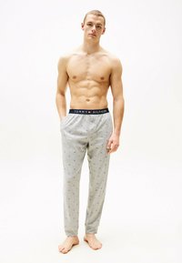 Pantalones de pijama grises con pequeños patrones geométricos, cinturilla elástica con el logo "TOMMY HILFIGER" y puños ajustados. El modelo está descalzo.