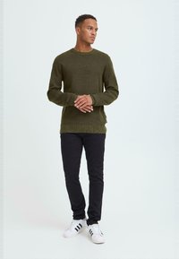 Maglione di colore verde oliva lavorato a maglia con scollo rotondo, tessuto testurizzato e polsini e orlo a costine, abbinato a jeans neri slim-fit e sneakers bianche.