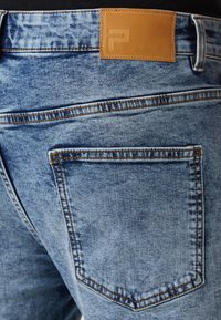 Vaqueros de mezclilla azul deslavado con una etiqueta de cuero en la cinturilla y dos bolsillos traseros con costuras contrastantes.