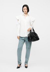 Femeie purtând o bluză albă cu mâneci cu volane, blugi albastru deschis, pantofi negri cu toc înalt, ținând o geantă mare neagră pe fundal simplu.
