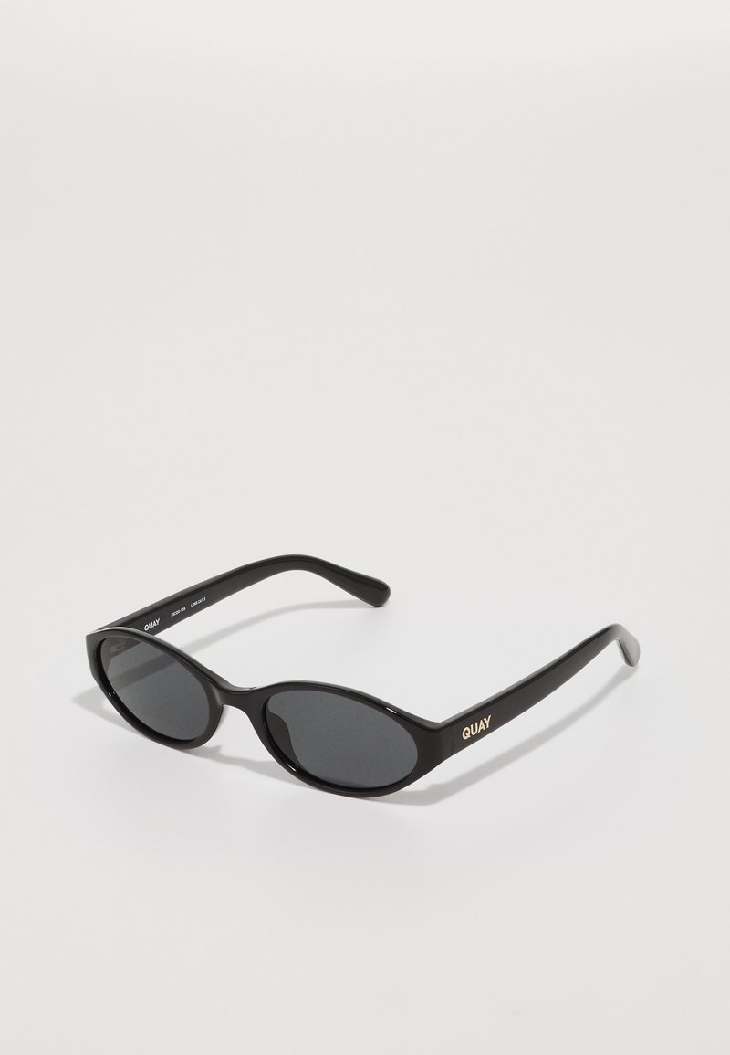 Lunettes de soleil noires avec des verres ronds, monture en plastique brillant et logo "QUAY" en doré sur le côté. Présente des verres teintés foncés.