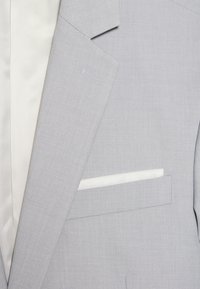 Hellegrauer Blazer aus fein strukturiertem Material. Mit einem weißen satinartigen Einstecktuch und einem beigen Innenfutter mit feinen Streifen.