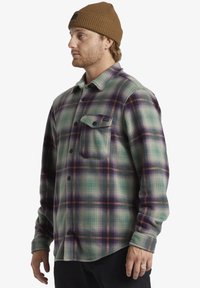 Groene geruite flanellen shirt met paarse en oranje accenten, voorzien van een borstzak, knopen en een relaxte pasvorm. Ggedragen met een bruine gebreide muts.