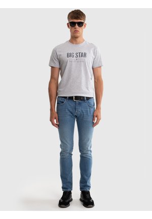 Mees, kandes musti päikeseprille, helehallit "BIG STAR" T-särki, siniseid pükse musta vöös ja musti tenniseid, seisab valgel taustal.