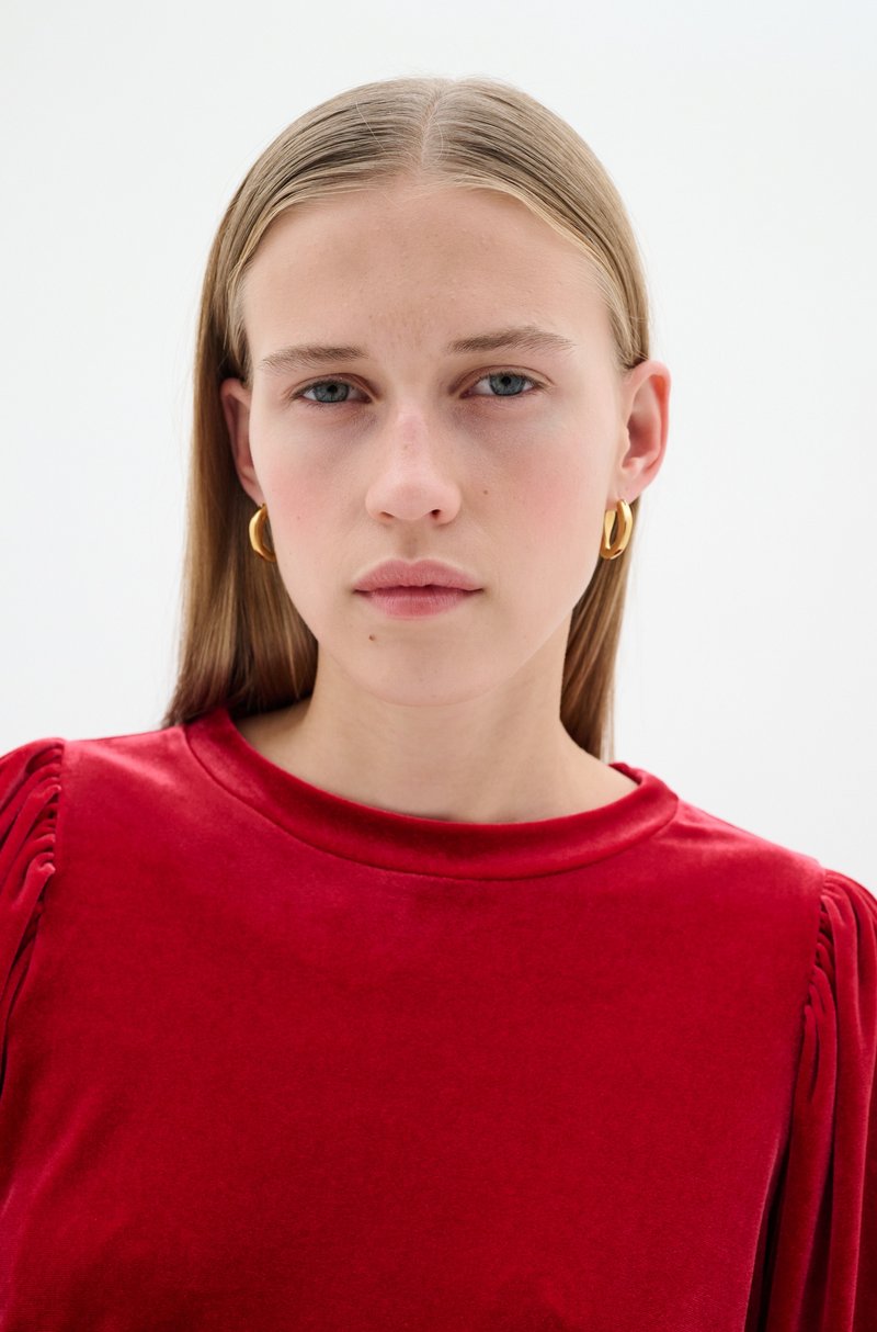 Rotes Samtoberteil mit gerafften Details an den Ärmeln, rundem Ausschnitt und glatter Textur. Das Model trägt goldene Ohrstecker und hat glattes, helles Haar.